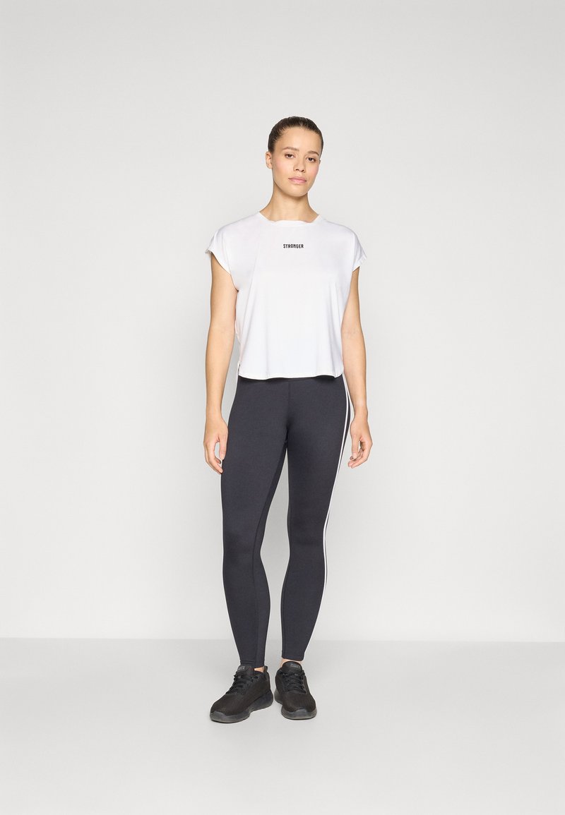 White cropped t-shirt with "STRONGER" text, paired with high-waisted black leggings featuring white side stripes. Black athletic shoes.