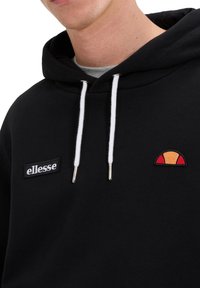 Sudadera negra con capucha y cordones blancos, con un pequeño logo negro de "ellesse" y un diseño colorido de medio sol en el pecho.