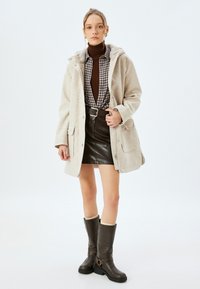 Chaqueta de mezcla de lana beige con capucha, llevada sobre un jersey marrón y una camisa de cuadros, combinada con una falda de cuero negra y botas marrones hasta la rodilla.