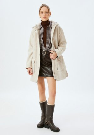Chaqueta de mezcla de lana beige con capucha, llevada sobre un jersey marrón y una camisa de cuadros, combinada con una falda de cuero negra y botas marrones hasta la rodilla.