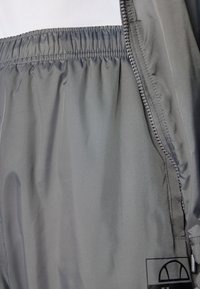 Gray nylon windbreaker with a smooth texture, elastic waistband, and zipper detail. Features a logo patch in the lower corner.