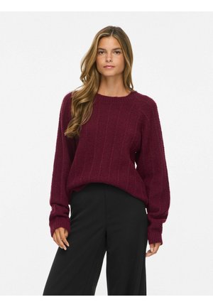 Femme aux longs cheveux ondulés portant un pull en maille rouge foncé et un pantalon large noir, debout devant un fond blanc uni.