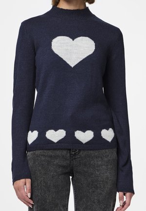 Marineblå sweater med et stort hvidt hjerte på brystet og en række mindre hvide hjerter langs kanten, båret med grå jeans.