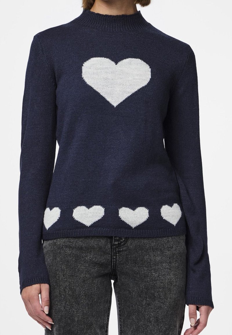 Pull bleu marine avec un grand cœur blanc sur la poitrine et une rangée de petits cœurs blancs le long de l'ourlet, porté avec un jean gris.