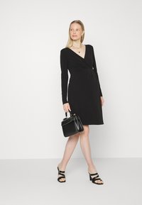 Vestido preto de mangas longas com design envolvente e decote em V. Tecido suave. Combinado com uma mala estruturada preta e sandálias pretas de salto bloco.