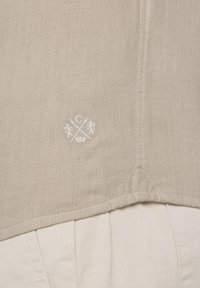 Linnen overhemd in een lichtbeige kleur, met een textuurstof, een geborduurd logo in het wit, en een geplooid ontwerp aan de onderkant.