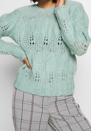 Femme portant un pull épais vert clair à motifs de feuilles et un pantalon à carreaux gris, posant avec une main sur la hanche et l'autre près du visage.