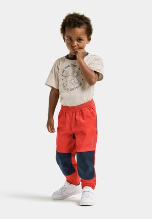 Røde og marineblå bukser med elastisk talje; beige t-shirt med et campingmotiv. Hvide sneakers fuldender outfittet. Barnet poserer eftertænksomt.