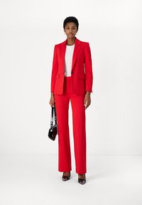 Rød skreddersydd dress med en tettsittende blazer og brede bukser, kombinert med en hvit topp. Tilbehøret inkluderer en liten svart håndveske og spisse hæler.