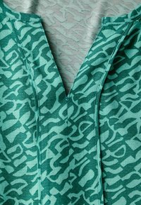 Haut en textile teal présentant un motif abstrait. Design en V avec une texture contrastante, tissu lisse et détails de couture visibles.