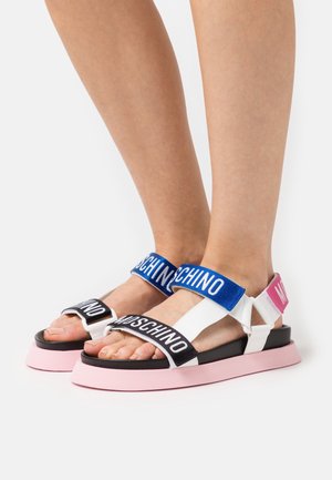 Nohy v bílých sandálech s černými, modrými a růžovými popruhy, černými podešvemi a silnými růžovými podrážkami na bílém pozadí.