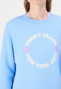Lys blå hettegenser med teksten "Tommy Jeans New York City" i en sirkel og to rosa hjerteikoner på brystet, båret av en person.