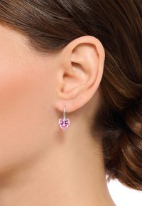 Orecchini a forma di cuore in un prezioso gemstone rosa, con montatura in argento e un semplice design ad anello, indossati su un orecchio liscio con capelli scuri.
