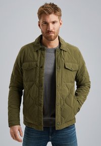 Olivgrüne gesteppte Jacke mit Druckknöpfen, Seitentaschen und Kragen, über einem grauen Sweater getragen, kombiniert mit blauen Jeans.