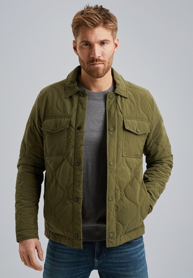 Olivgrüne gesteppte Jacke mit Druckknöpfen, Seitentaschen und Kragen, über einem grauen Sweater getragen, kombiniert mit blauen Jeans.