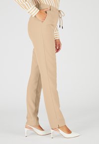 Beige op maat gemaakte broek met voorzak, taille met koord en rechte pijpen. Gecombineerd met witte hoge hakken.