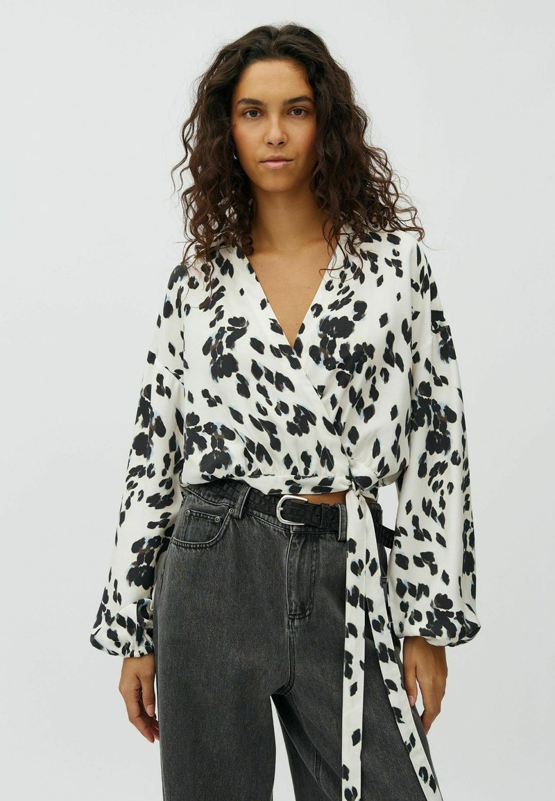 Femme aux cheveux bouclés portant un chemisier portefeuille blanc à manches longues avec des taches florales noires et un jean noir délavé taille haute.