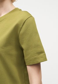 Άτομο που φοράει ελιάς πράσινο κοντομάνικο με στρογγυλή λαιμόκοψη, εμφανιζόμενο από τον ώμο έως το μέσο του βραχίονα σε απλό φωτεινό φόντο.