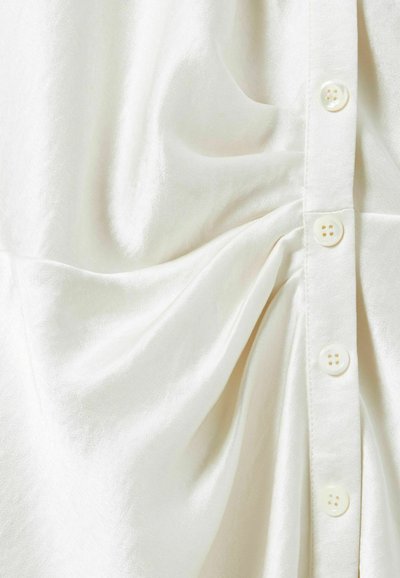 Blusa bianca in tessuto liscio, con patta a bottoni e dettagli arricciati su un lato. Texture morbida con una leggera lucentezza.