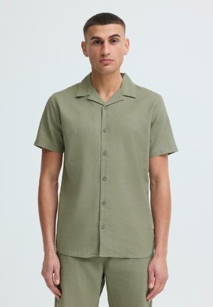 Mann trägt ein kurzärmeliges, olivgrünes Hemd mit Knöpfen und passende Hose, steht vor einem schlichten, hellen Hintergrund.