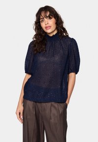 Tengerészkék áttetsző blúz texturált anyagból, magas nyakkal és puffos rövid ujjakkal, barna pliszírozott nadrággal párosítva.