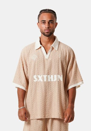 Man met gevlochten haar draagt een losse beige gebreide trui en bijpassende korte broek met een witte kraag en mouwranden tegen een effen achtergrond.