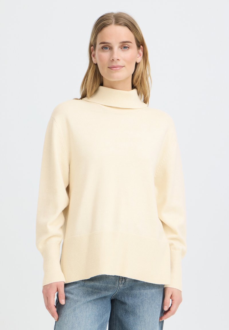 Femme portant un pull à col roulé crème à manches longues et un jean bleu, debout devant un fond clair uni.