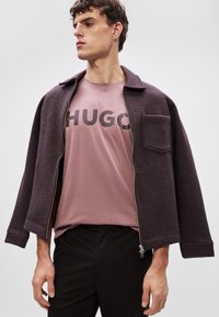 Tmavohnedá zipsová bunda s golierom, nosená cez svetloružové tričko s tučným čiernym písmom. Čierne nohavice dopĺňajú celkový outfit.