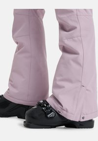 Lichtpaarse skibroek met een gladde textuur, taps toelopend ontwerp en drukknoopsluiting bij de enkel, gecombineerd met zwarte skijschoenen met hardware-accenten.