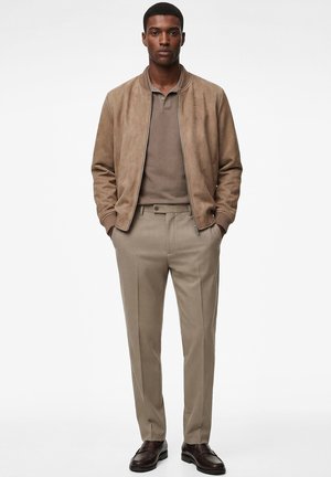 Hombre de pie de frente, vistiendo una chaqueta bomber de ante color beige, camisa polo marrón, pantalones beige y zapatos de cuero marrón oscuro.