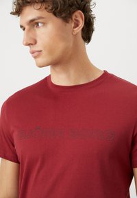 Tricou roșu din bumbac, cu guler rotund și mâneci scurte, având textul "BJÖRN BORG" imprimat în gri închis pe piept.