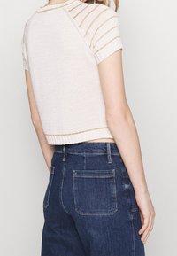 Suéter cropped de punto en beige claro con rayas metálicas doradas en la parte superior. Combinado con jeans de mezclilla oscuros de tiro alto con bolsillos traseros.