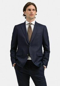 Marinblå kostymjacka med en strukturerad design, enkel knappstängning och två framfickor. Vit skjorta och slips med bruna prickar.