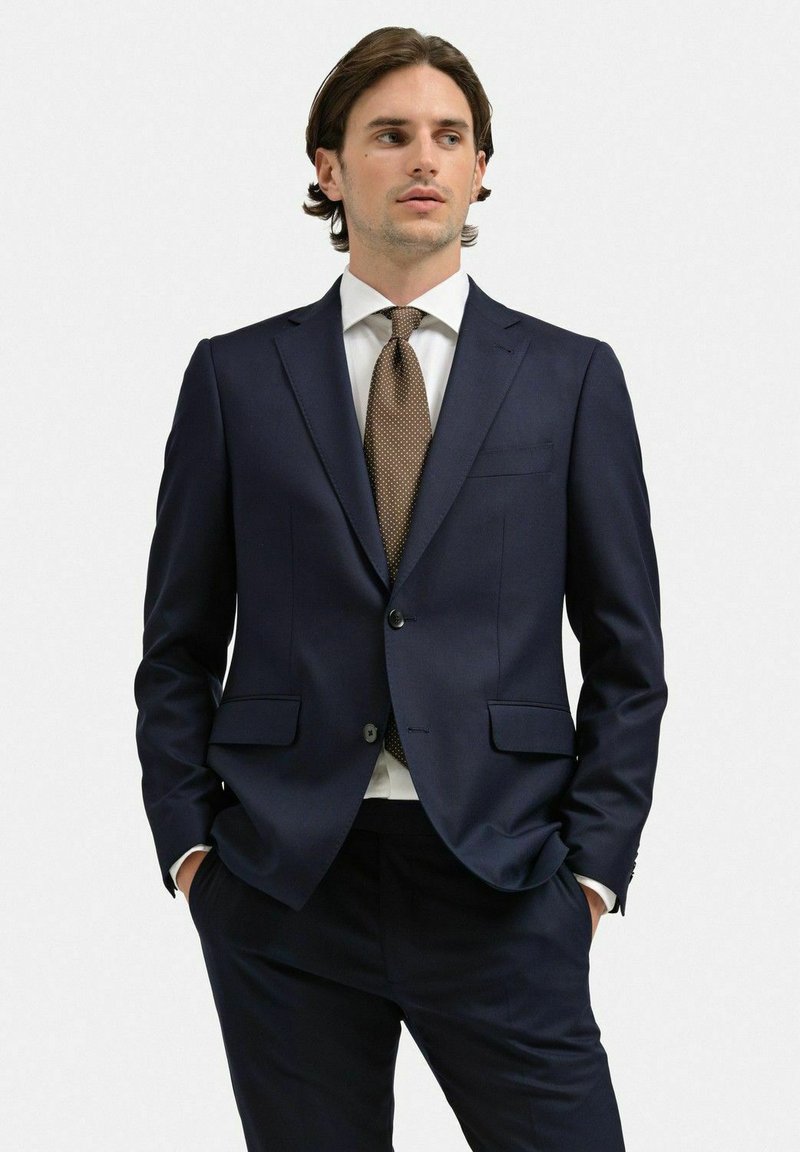 Marinblå kostymjacka med en strukturerad design, enkel knappstängning och två framfickor. Vit skjorta och slips med bruna prickar.