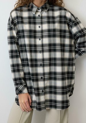Kobieta w oversize czarno-biało-szarej kratowanej koszuli zapinanej na guziki i beżowych spodniach na jednolitym białym tle.