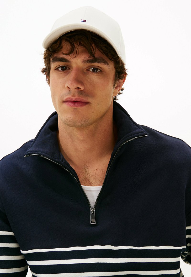 Suéter navy con rayas horizontales blancas y cuello con cremallera; llevado sobre una camiseta blanca. Gorra blanca en la cabeza.