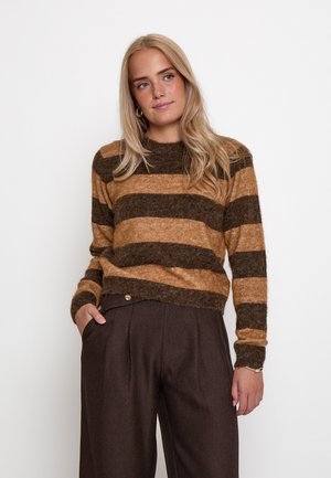 Blonde vrouw die een bruin- en beige gestreepte trui en donkerbruine wijde broek draagt, staand met één hand in de zak tegen een witte achtergrond.