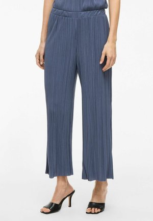 Wijde, geplooide pantalons in een donkere blauwe stof, met een rekbare tailleband en zijsplitsen aan de zoom, gedragen met zwarte open teen hakken.