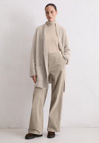 Beige, fluffy cardigan met open voorkant, gedragen over een beige coltrui, gecombineerd met wijdvallende beige broek en donkerbruine schoenen. Textuur details van de stof.