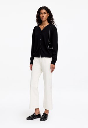 Vrouw draagt een zwarte gebuttonneerde cardigan, witte broek met wijde pijpen, zwarte loafers en draagt een kleine zwarte schoudertas, staand op een witte achtergrond.