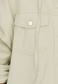 Gros plan sur la poche d'une veste en tissu beige avec un rabat sécurisé par un bouton rond argenté et un cordon attaché qui pend.