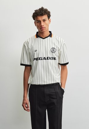 Giovane uomo con capelli ricci che indossa una maglia sportiva Pegador bianca a righe sottili e pantaloni neri, in piedi davanti a uno sfondo chiaro e semplice.