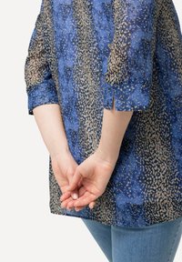 Blusa blu e beige a fantasia con maniche a tre quarti, caratterizzata da un dettaglio con orlo spaccato e un tessuto leggero e trasparente.