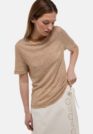 Femme ajustant une jupe portefeuille beige avec broderies florales, portant un T-shirt rayé marron et blanc à manches courtes sur fond blanc.