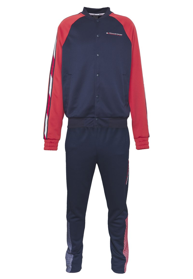 Tuta blu navy con maniche raglan rosse, giacca con bottoni e polsini elastici. I pantaloni presentano pannelli laterali con un motivo geometrico rosso e blu.