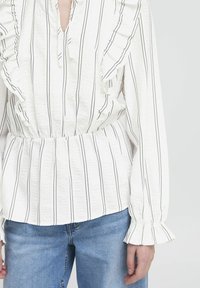 Blusa bianca con strisce verticali nere, dettagli arricciati lungo il colletto e i polsini, vita arricciata, abbinata a jeans in denim blu.