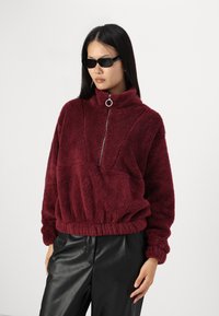 Burgunderfarget fleece-pull med høy krage, sølvglidelås og elastisk linning. Teksturert overflate med synlige sømmer og avslappet passform.