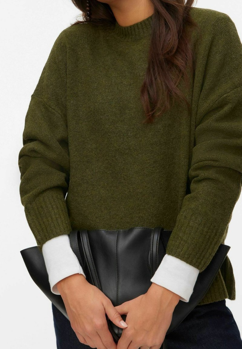 Personne portant un pull en tricot vert olive avec des manches blanches apparentes, tenant une grande pochette en cuir noir au niveau de la taille.