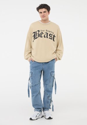 Felpa beige con testo grafico nero "Los Angeles Beast"; jeans cargo blu con molte tasche; sneakers bianche e grigie.