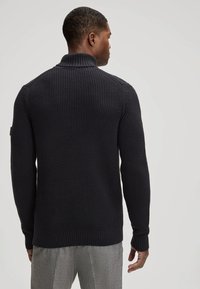 Pull en maille bleu marine foncé avec un col montant, surface texturée et poignets et ourlet côtelés, porté avec un pantalon ajusté gris clair.